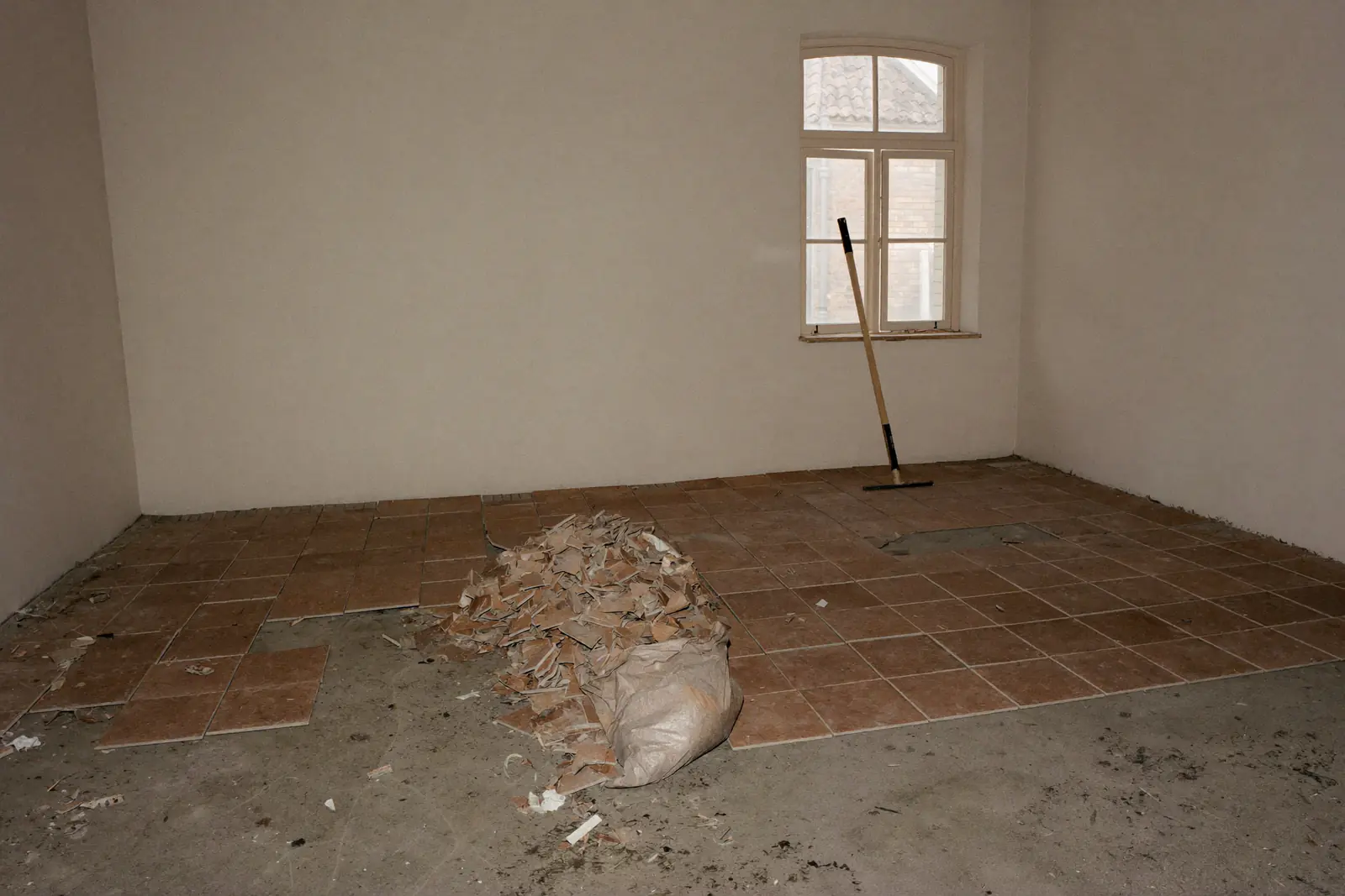Lege voorkamer van een oud bakstenen huis waar een deel van de bruine tegelvloer al is verwijderd en de grijze dekvloer zichtbaar is, met een zak tegelpuin en een schraper bij het raam