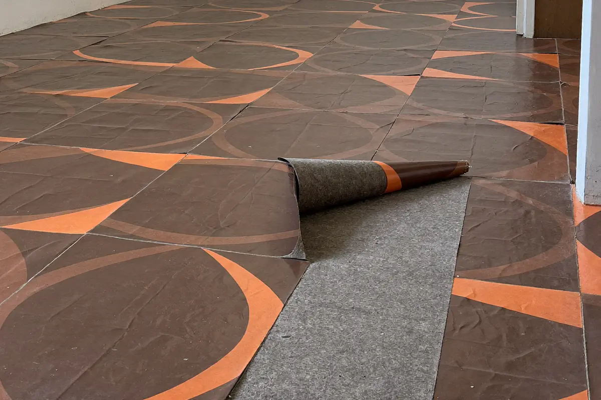 Oud bruin vloerzeil met oranje cirkelpatroon in een lege kamer, met een opgerolde hoek waaronder een grijze viltlaag zit